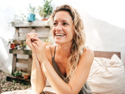 Joyful woman with wavy blonde hair smiling, seated outdoors beside a wooden pallet garden shelf with plants.