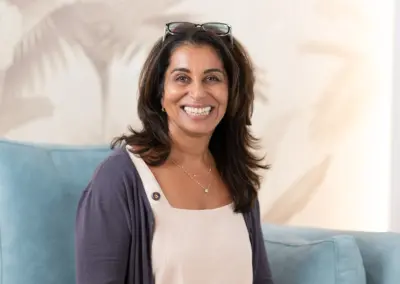 Smiling woman seated on a light-blue sofa wearing a white top and navy cardigan, sunglasses on her head.