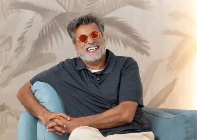 Smiling older man with gray hair and orange round sunglasses sits on a light-blue chair, arms resting, palm-leaf mural backdrop behind him.