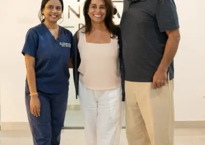 Three smiling adults stand together in a bright clinic lobby, with a medical professional in blue scrubs on the left, a woman in white in the center, and a man in a dark polo with orange sunglasses on the right, in front of a logo sign.